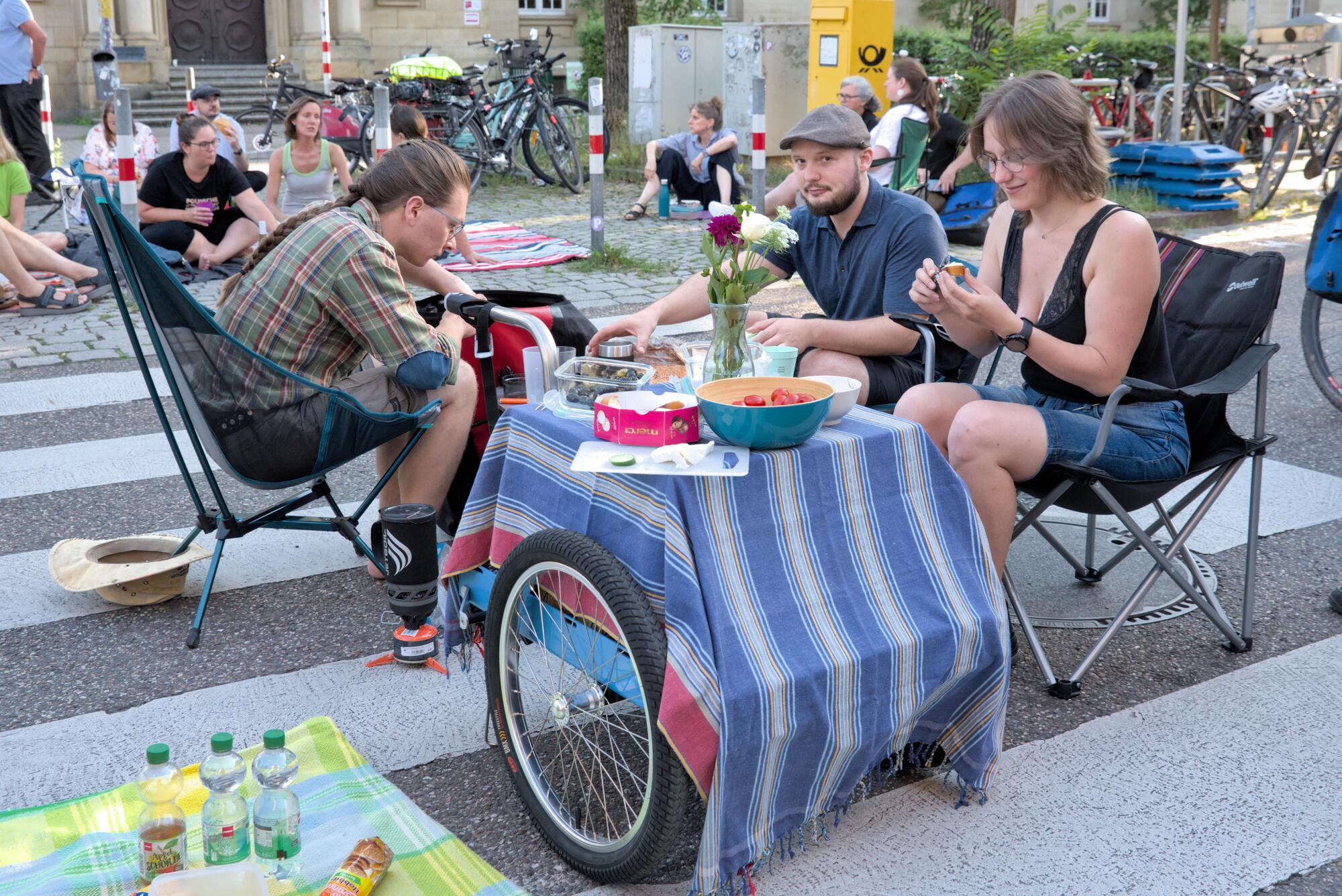 Personen sitzen bei einem Picknick auf einem Zebrastreifen auf der Sophienstraße am Gutenbergplatz. Sie sitzen auf Faltstühlen um einen kleinen Tisch herum, der aus einem niedrigen Fahrradanhänger besteht.