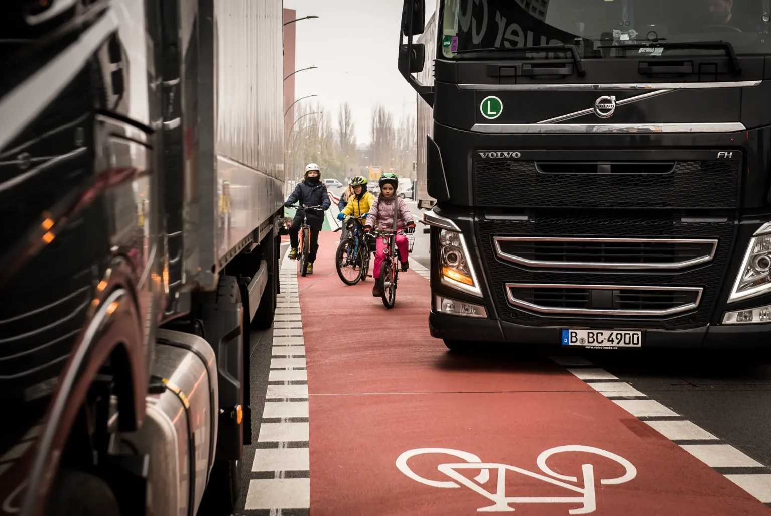 Kinder und Erwachsene fahren mit Fahrrädern auf einem Radfahrstreifen in Mittellage zwischen Lastkraftwagen hindurch. Einer der beiden setzt zum Fahrstreifenwechsel über den Radfahrstreifen hinweg an, während die Radfahrenden im toten Winkel sind.